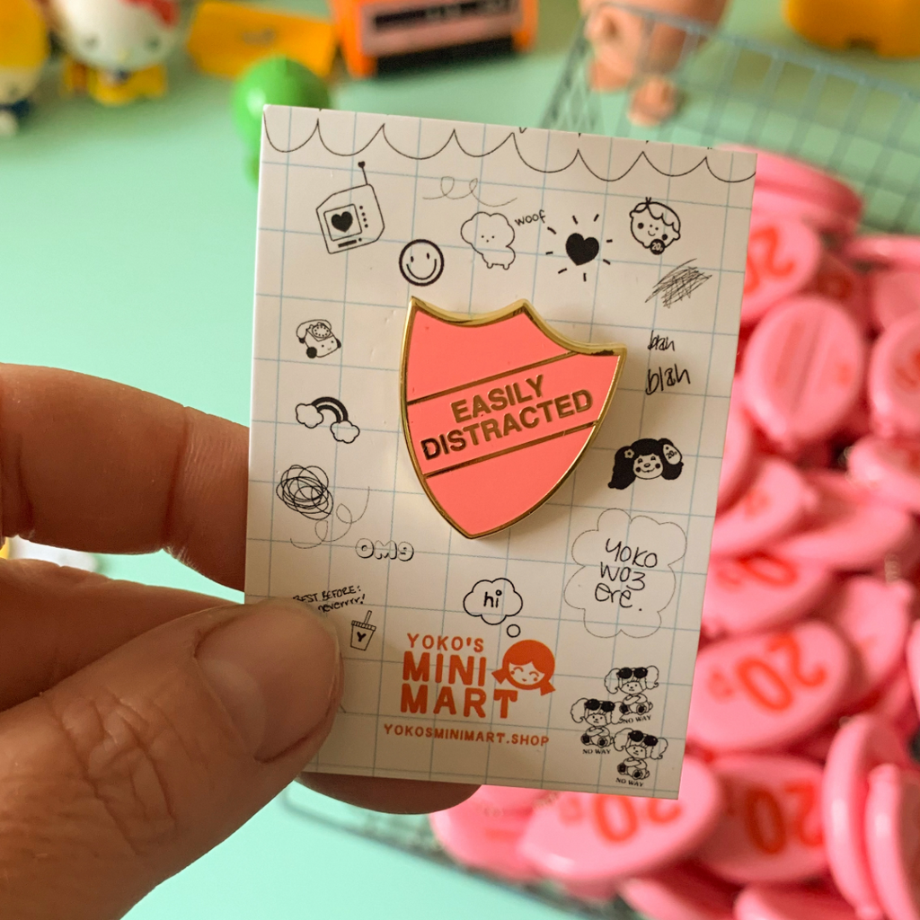 Pink enamel pin with 'Easily Distracted' text on a card, held by a hand with a colorful background.